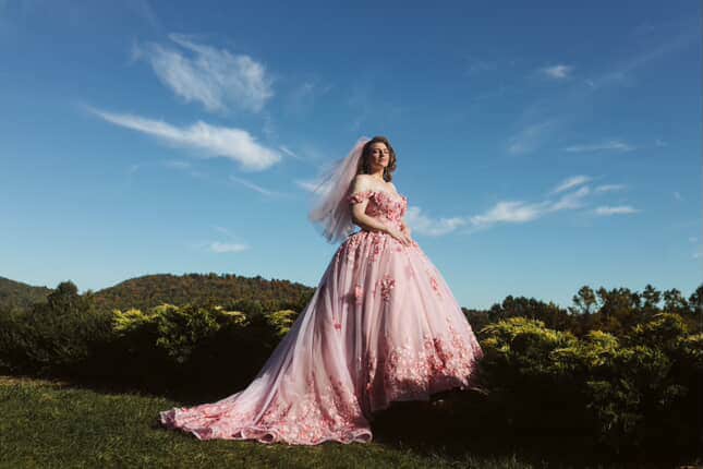Bride posing on a hilltop in the custom pink ball gown, off-shoulder bodice and sweeping floral train