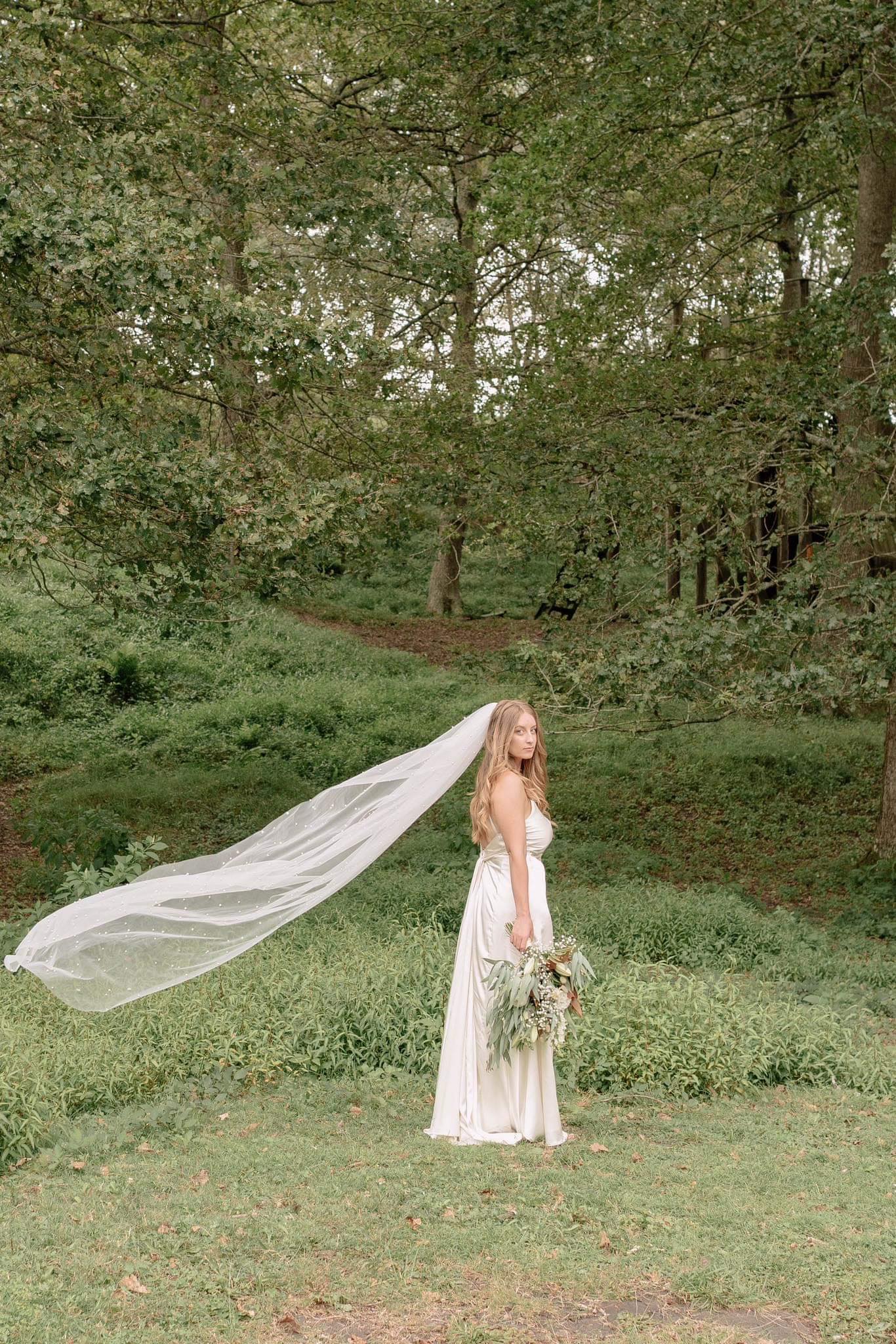 Bride in flowing ivory silk custom made dress with long sheer veil and greenery bouquet outdoors – Art Kollocsion Auckland
