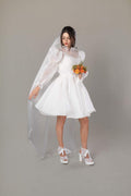 Bride in white mini wedding dress with embroidered tulle veil, sheer puff sleeves and orange bouquet, front view