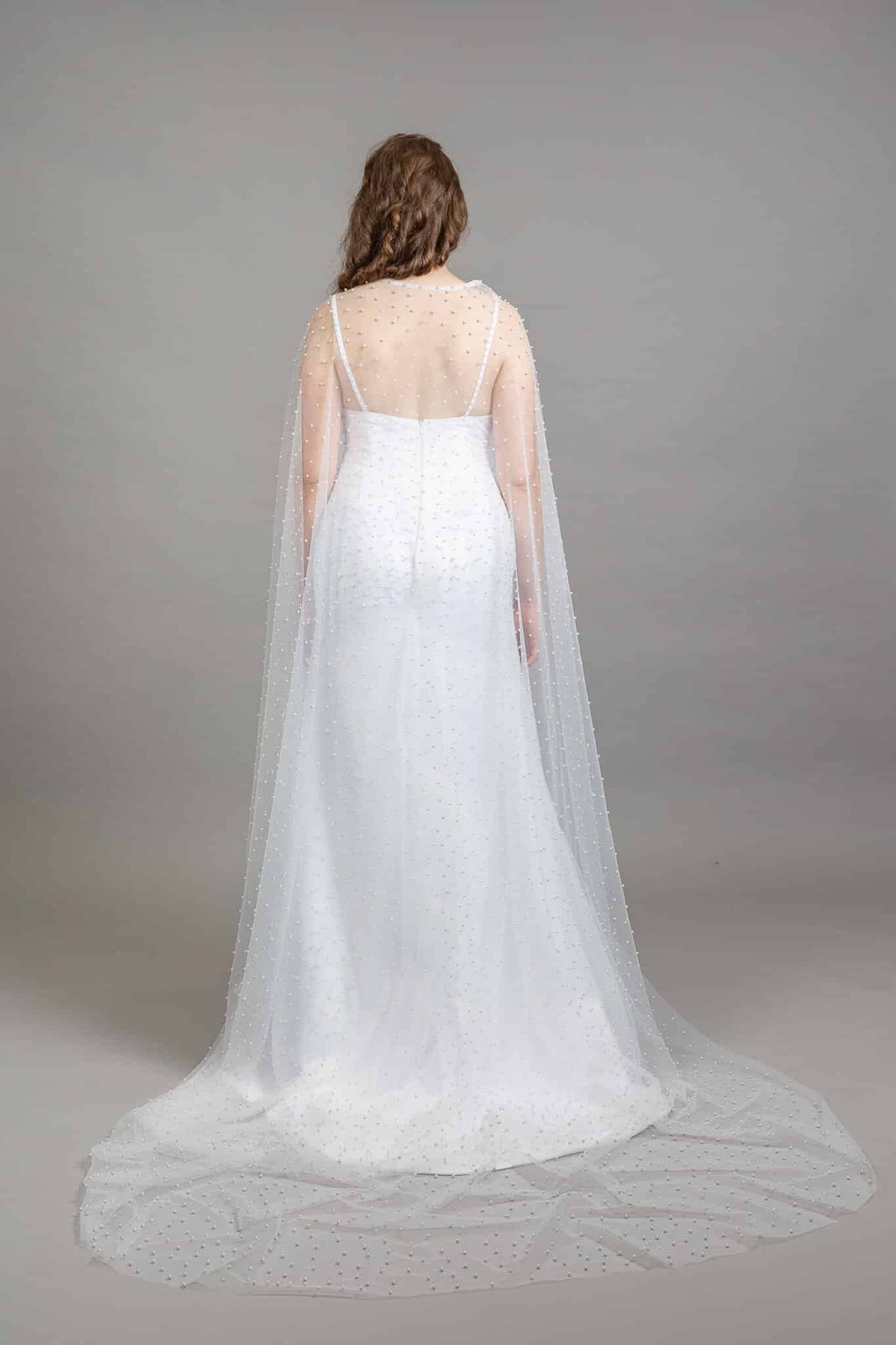 Back view of white sheath wedding gown with open back and sheer pearl tulle cape flowing into a long train