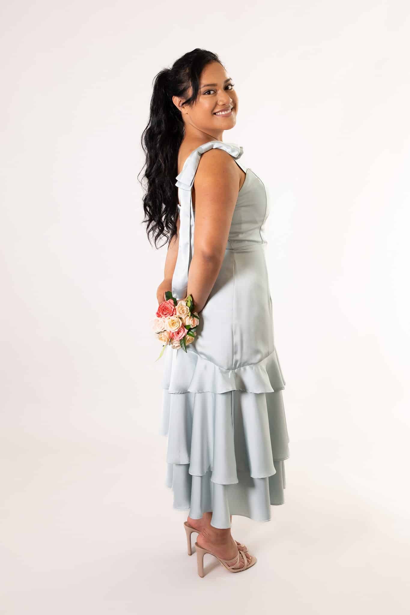 Pale blue satin midi dress, layered ruffle hem, bow-shoulder detail, side profile with bouquet