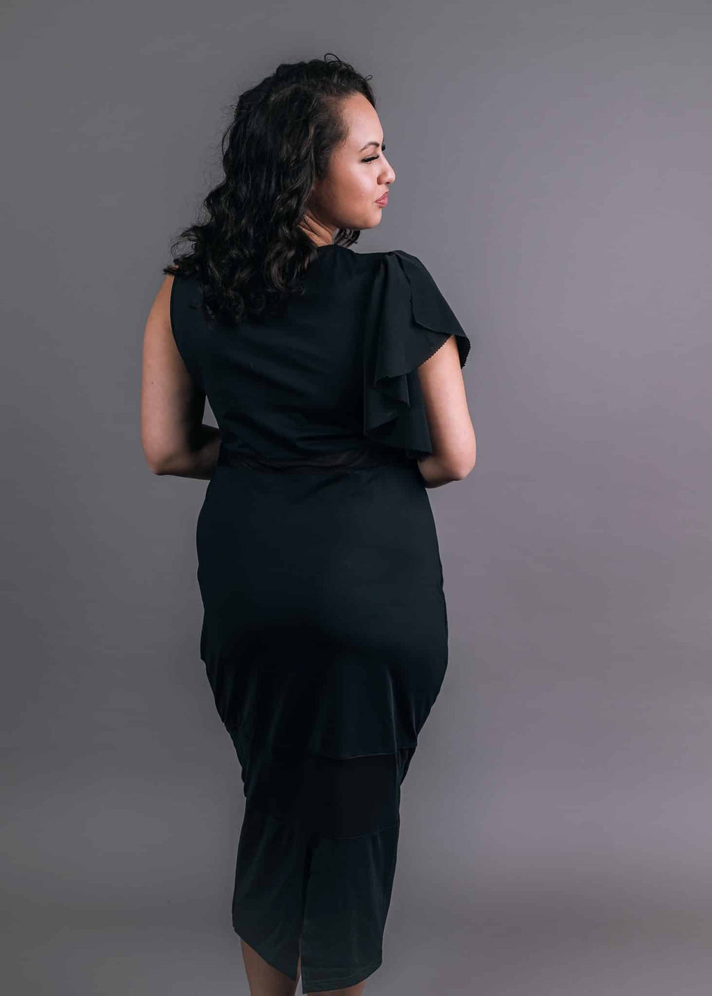 Back view showing a ruffled black attachable sleeve cascading from one shoulder of a black bodycon midi dress
