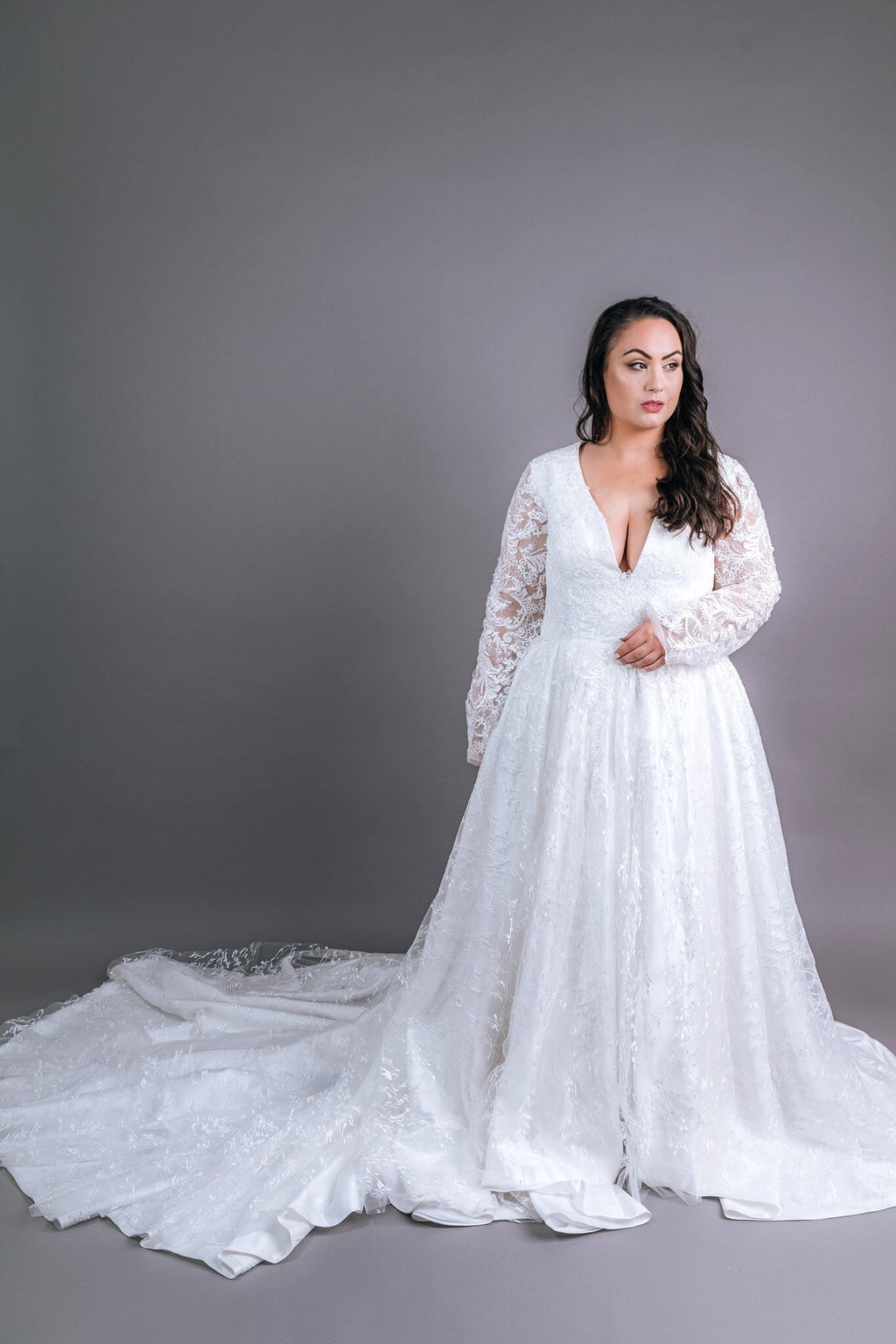Model in affordable plunge neckline wedding gown, long lace sleeves and flowing lace train, front three-quarter pose