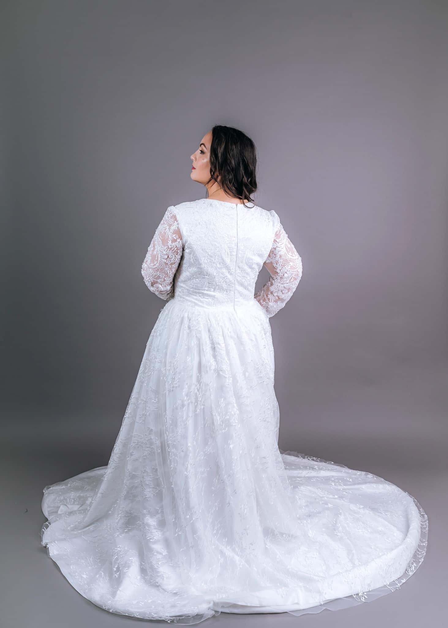 White long-sleeve lace wedding gown from behind, showing zip-back bodice and full tulle train pooling around model