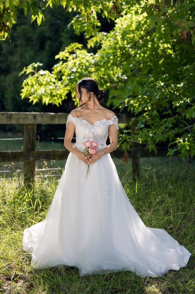 Bride holding pink bouquet, showing intricate lace appliques on off-shoulder bodice