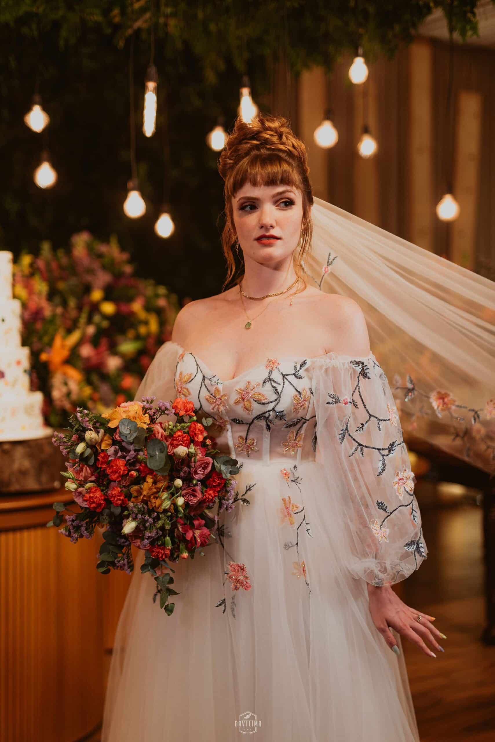 Bride wearing off-shoulder non-traditional wedding dress with colorful floral embroidery and trailing veil, holding bouquet